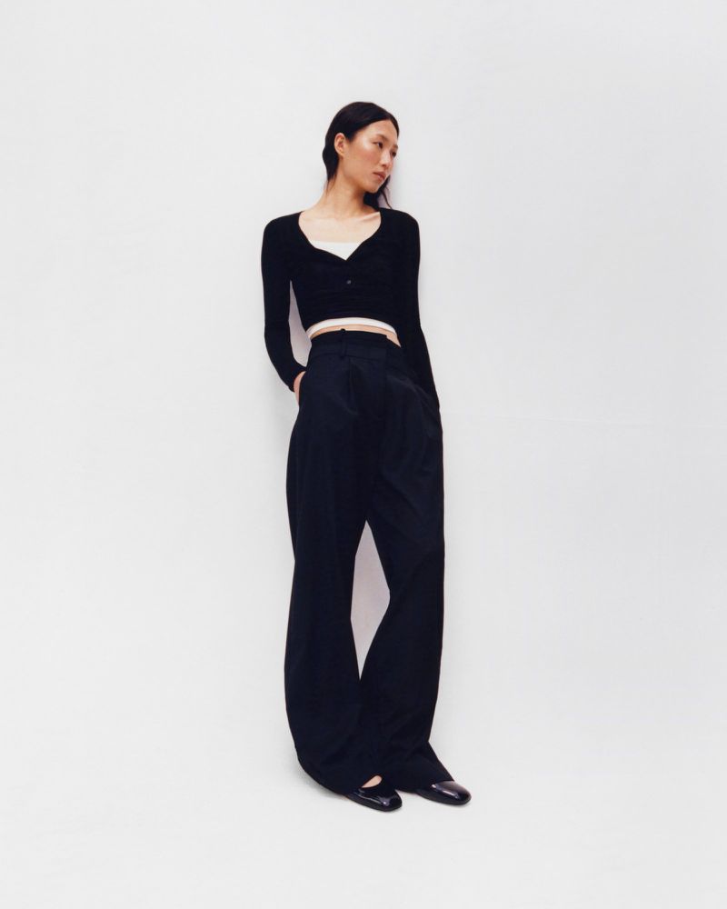 A slender Asian woman poses in a black cardigan and wide-leg trousers against a bright white backdrop in a full-length fashion shot.