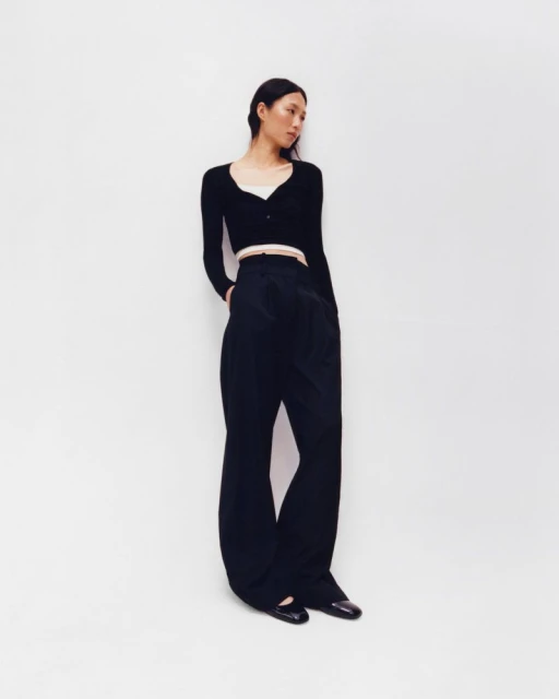 A slender Asian woman poses in a black cardigan and wide-leg trousers against a bright white backdrop in a full-length fashion shot.