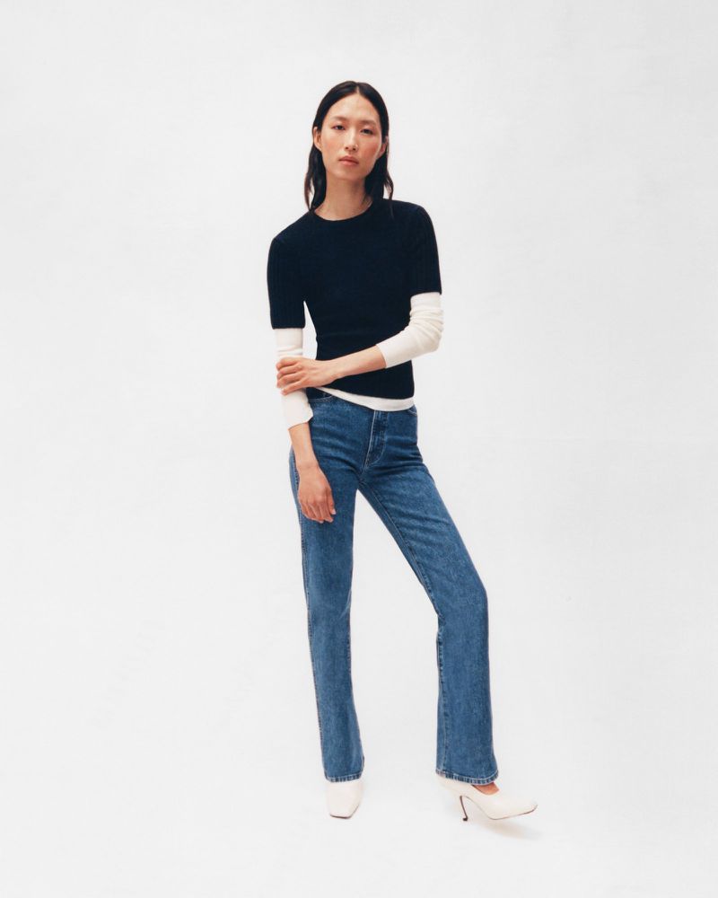 Full shot of a slender Asian woman wearing a navy short-sleeve sweater and jeans in a brightly lit photo studio on a white backdrop.