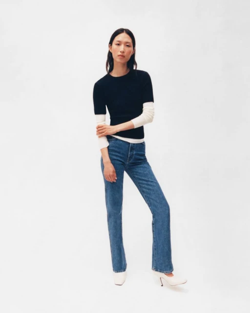 Full shot of a slender Asian woman wearing a navy short-sleeve sweater and jeans in a brightly lit photo studio on a white backdrop.