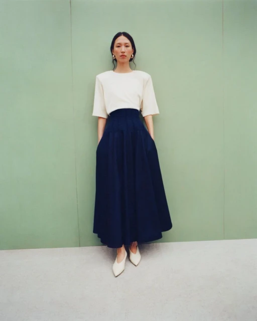 Full-shot fashion portrait of a slender adult woman wearing a cream top and navy skirt standing against a green wall in a photo studio.