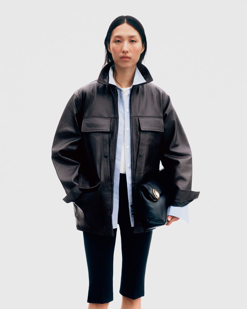 A woman stands in a studio wearing a brown leather jacket, white shirt, and black culottes, holding a black leather purse.
