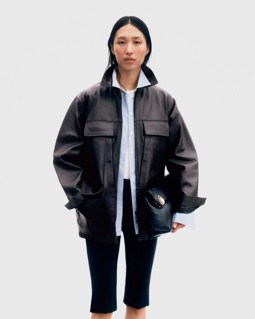 A woman stands in a studio wearing a brown leather jacket, white shirt, and black culottes, holding a black leather purse.