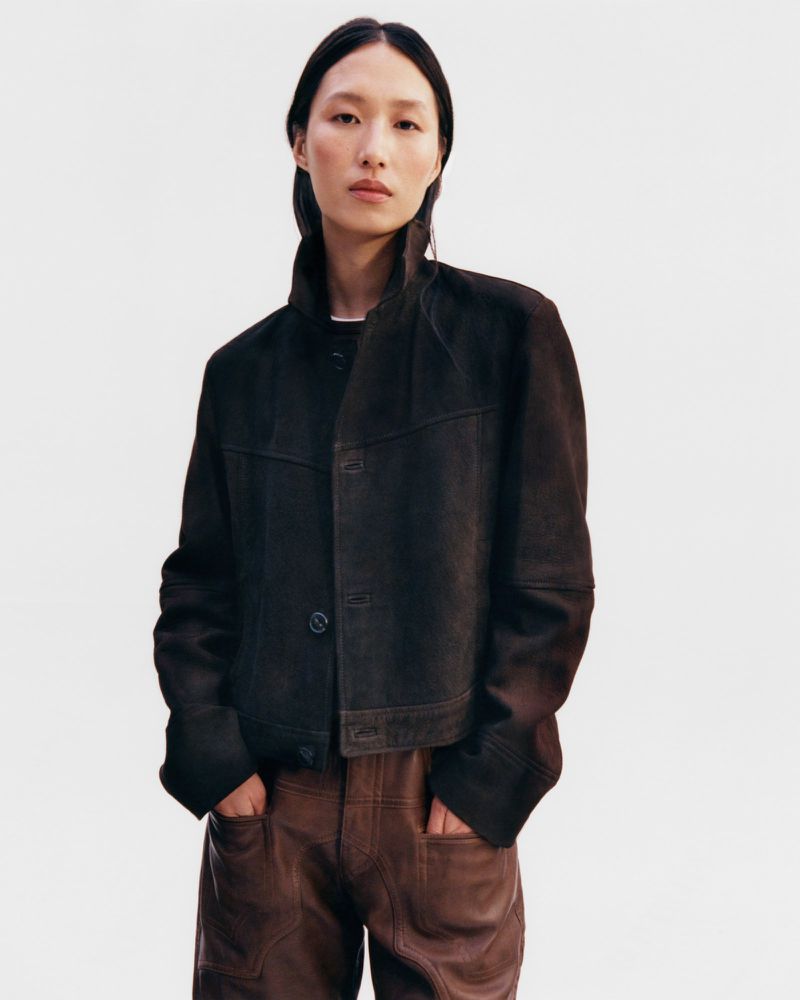 An Asian female model poses indoors wearing a dark brown suede jacket and brown leather pants against a neutral studio background.