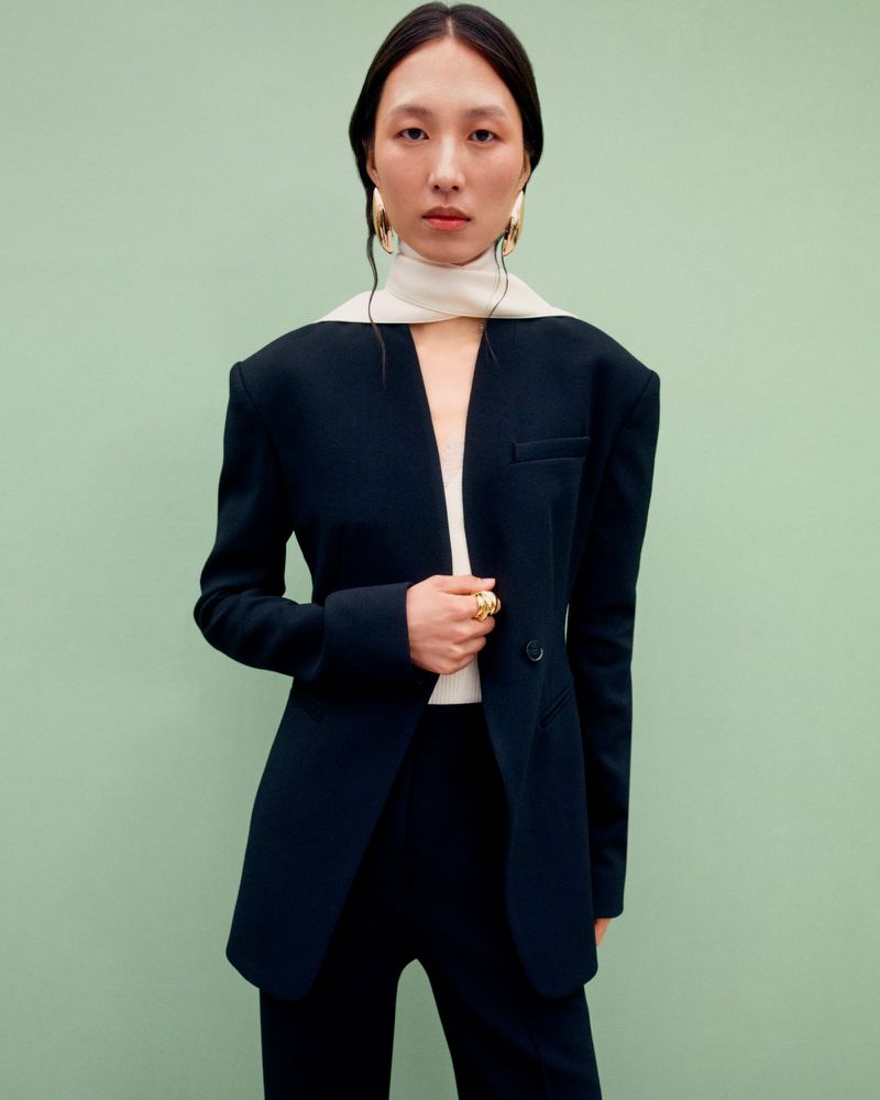 Elegant Asian woman in tailored black pantsuit with statement gold jewelry poses against a serene pastel green background in a fashion portrait.