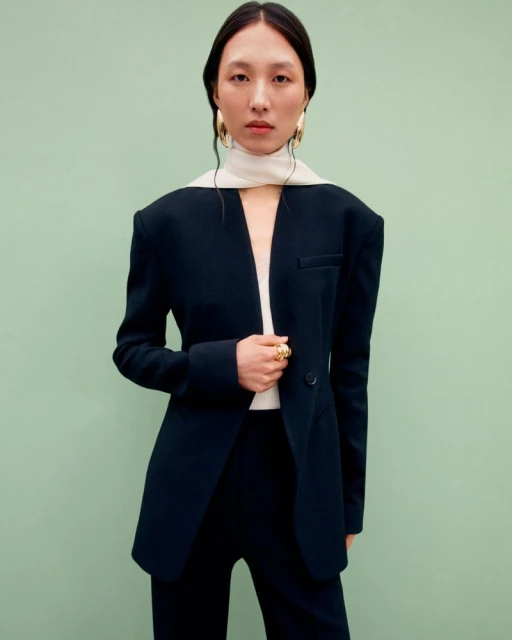 Elegant Asian woman in tailored black pantsuit with statement gold jewelry poses against a serene pastel green background in a fashion portrait.