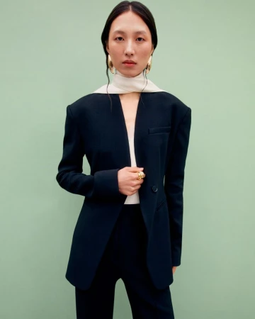 Elegant Asian woman in tailored black pantsuit with statement gold jewelry poses against a serene pastel green background in a fashion portrait.