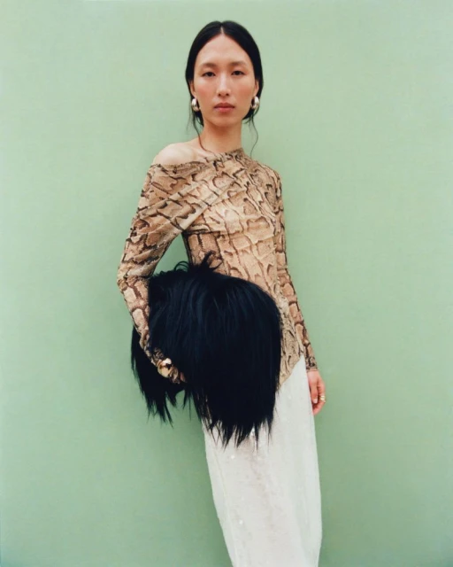 Portrait of an Asian woman in a snakeskin top and white skirt, holding a black faux fur muff against a light green background.