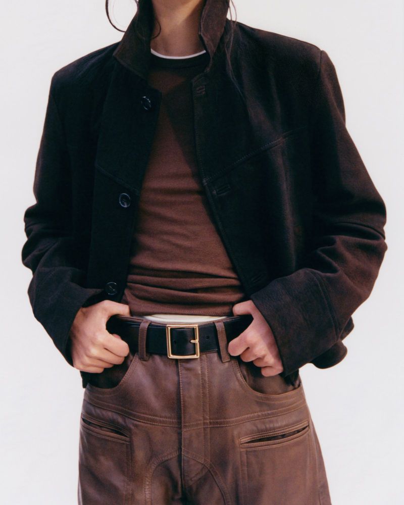 Person in brown leather pants and a brown jacket posing in front of a neutral background in a fashion studio shot. 