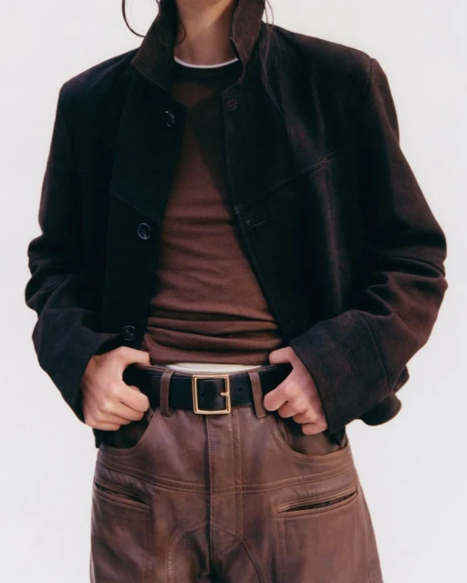 Person in brown leather pants and a brown jacket posing in front of a neutral background in a fashion studio shot. 