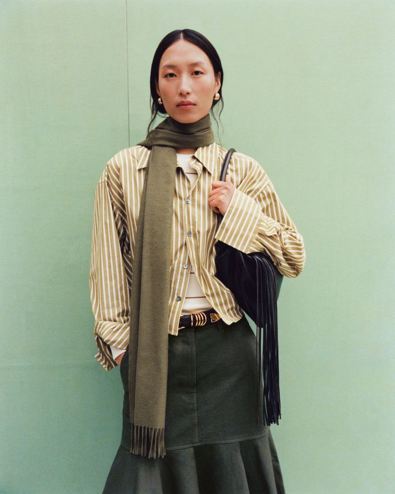 Fashion portrait of an Asian woman wearing a striped shirt, green skirt, and scarf, posing against a light green backdrop in a studio.
