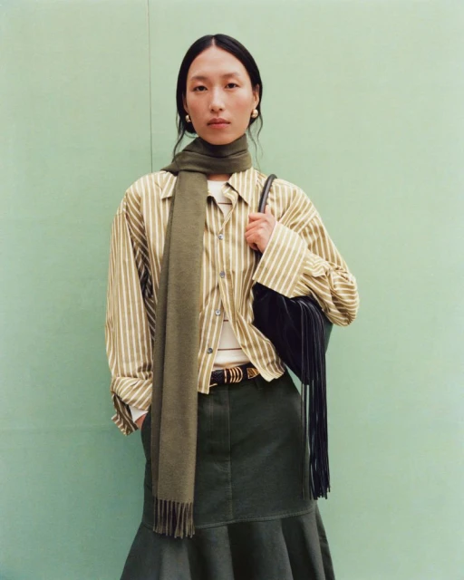 Fashion portrait of an Asian woman wearing a striped shirt, green skirt, and scarf, posing against a light green backdrop in a studio.