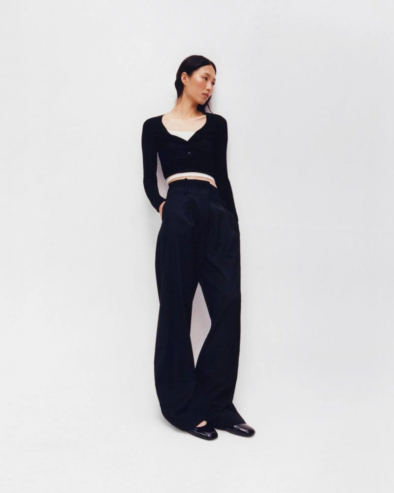 Model wearing a black cardigan and high-waisted trousers standing against a white backdrop.