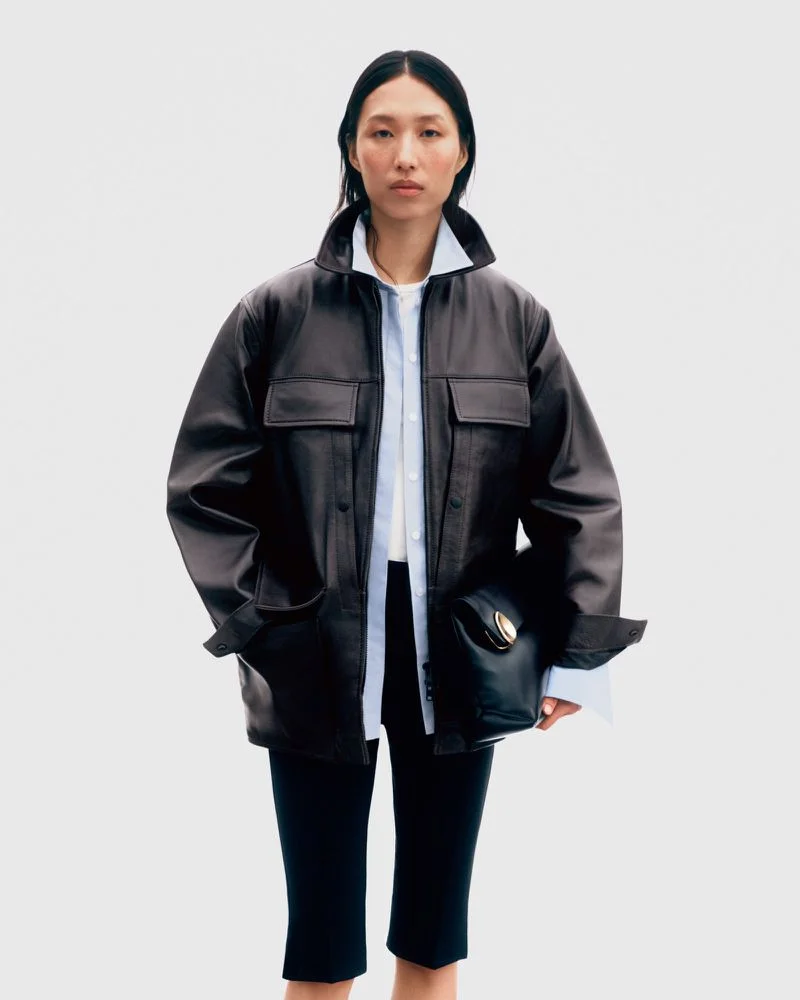 Model wearing a black leather jacket over a light shirt holding a black handbag against a white background