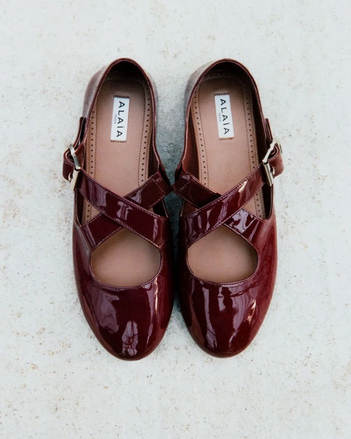 Top-down view of a pair of glossy burgundy patent leather Mary Jane flats with crisscross straps and silver buckles on a light background.