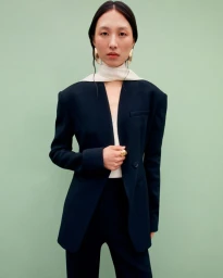 Model wearing a structured dark blazer and white scarf against a light green seamless studio backdrop.