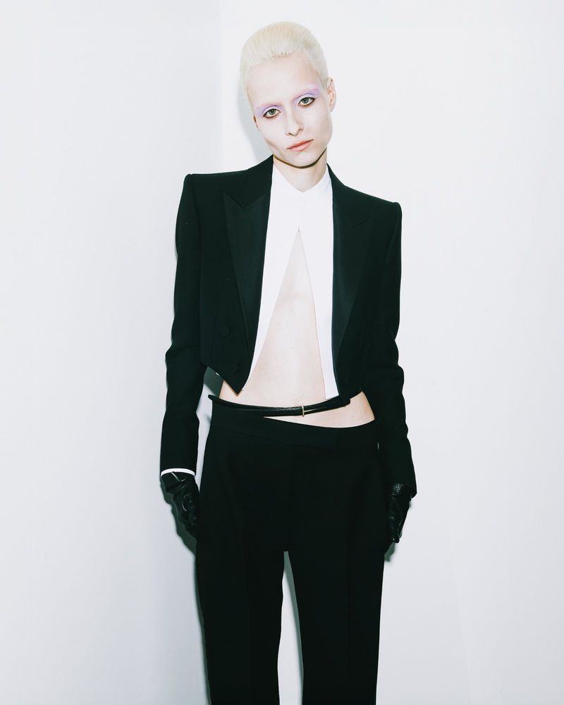 A pale-skinned model with cropped platinum hair poses in a black cropped blazer and matching trousers against a white backdrop in a studio.