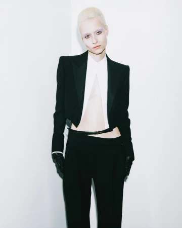 A pale-skinned model with cropped platinum hair poses in a black cropped blazer and matching trousers against a white backdrop in a studio.