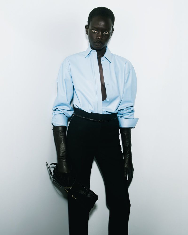Fashion shot of a Black adult female in a light blue button-down shirt, black pants, and black leather gloves, holding a black purse.