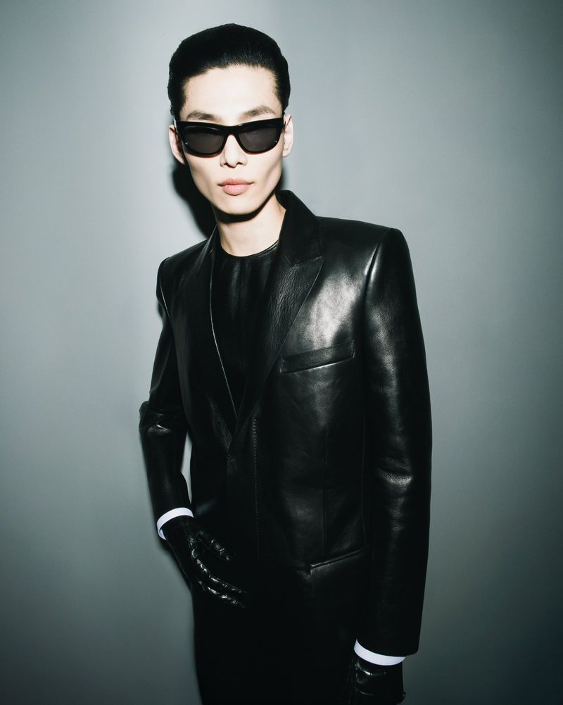 Portrait of a man in a black leather suit and sunglasses posing against a gray background with studio lighting; fashion editorial shot.