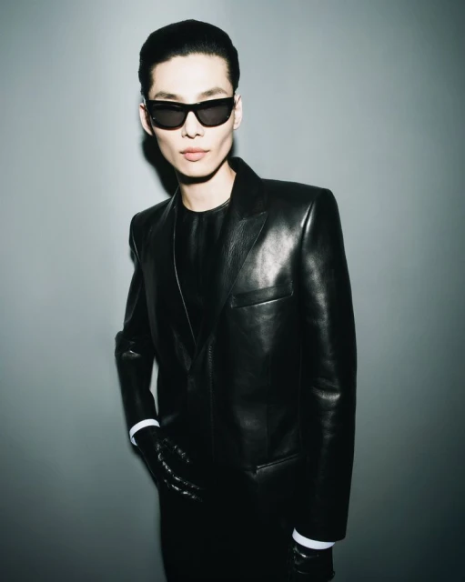 Portrait of a man in a black leather suit and sunglasses posing against a gray background with studio lighting; fashion editorial shot.