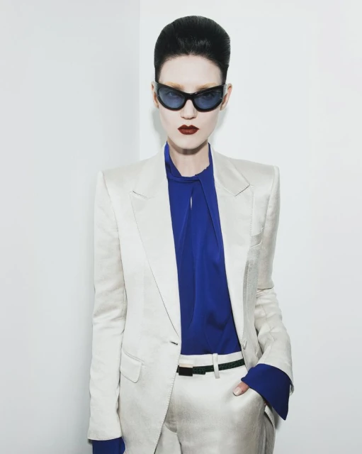A stylish woman with pale skin and dark hair poses in a white pantsuit with a blue top and dark sunglasses in a studio.