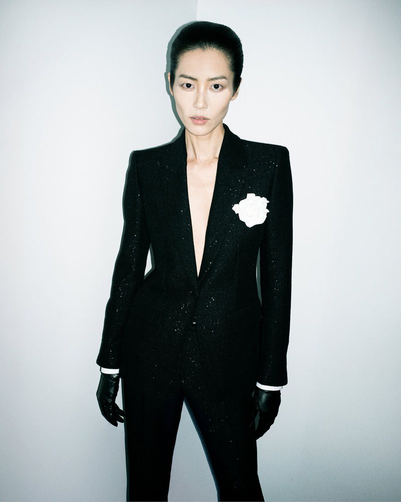 Elegant woman in a black, glittering suit with a white flower boutonniere, wearing black gloves, posing against a neutral wall backdrop.