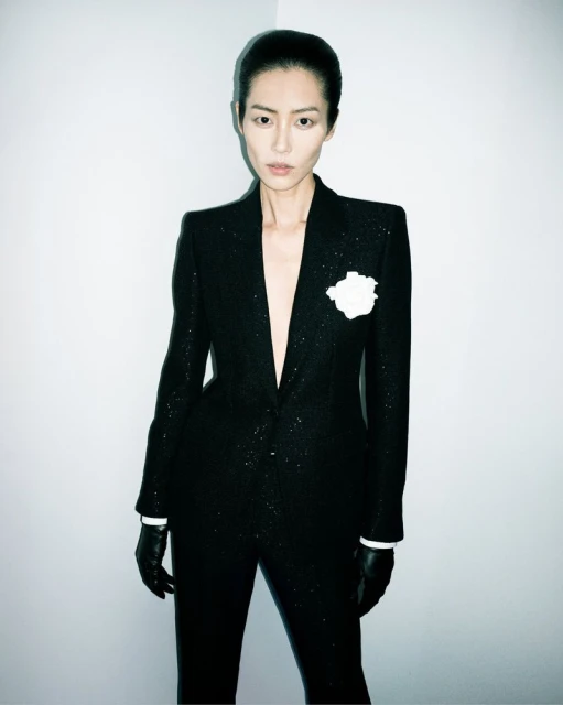 Elegant woman in a black, glittering suit with a white flower boutonniere, wearing black gloves, posing against a neutral wall backdrop.