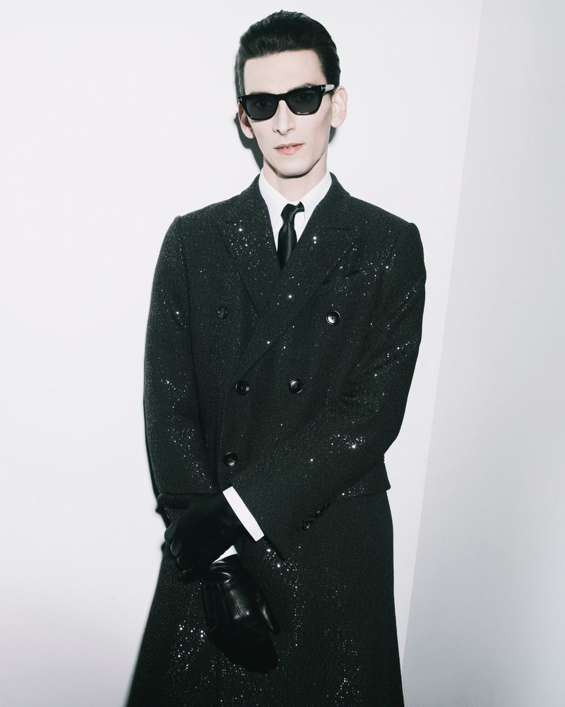Portrait of a stylish adult male wearing sunglasses, a glittering black overcoat, black gloves, and a white dress shirt against a white wall.
