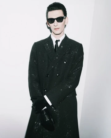 Portrait of a stylish adult male wearing sunglasses, a glittering black overcoat, black gloves, and a white dress shirt against a white wall.