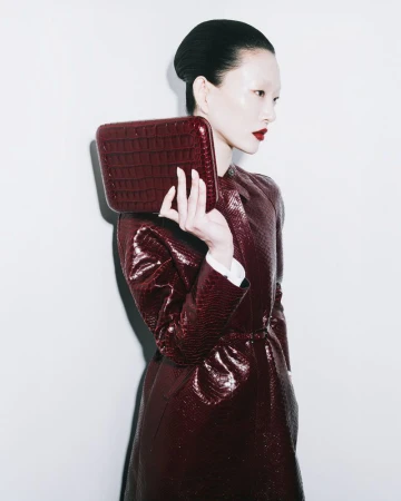 Fashion photo of an adult woman holding a structured box clutch. She wears a matching burgundy coat and dark red lipstick.
