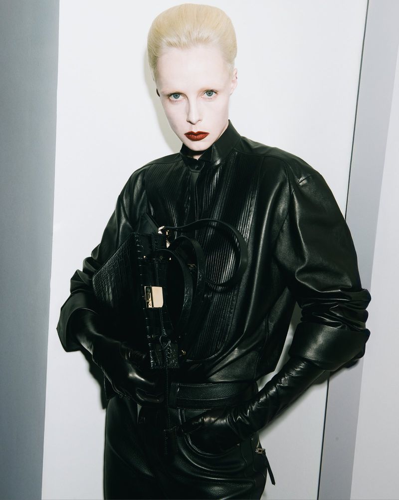 A fair-skinned model with platinum blonde hair in a black leather outfit poses indoors with a black leather purse.