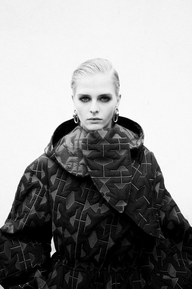 Monochrome portrait of adult female in patterned hooded coat wearing hoop earrings against off-white seamless backdrop in studio.