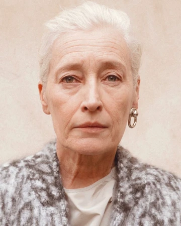 Close-up portrait of an older woman with white hair wearing a patterned grey jacket and silver earring against a neutral wall.