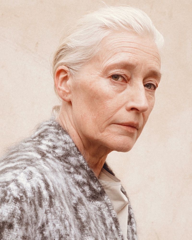 Close-up portrait of an adult woman with white hair in a light brown and gray jacket against a neutral-toned background.