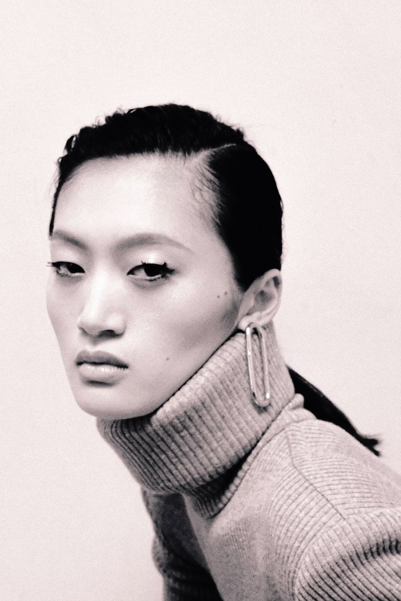 A close-up black and white portrait of an Asian woman with short dark hair, wearing a gray turtleneck and a silver earring.