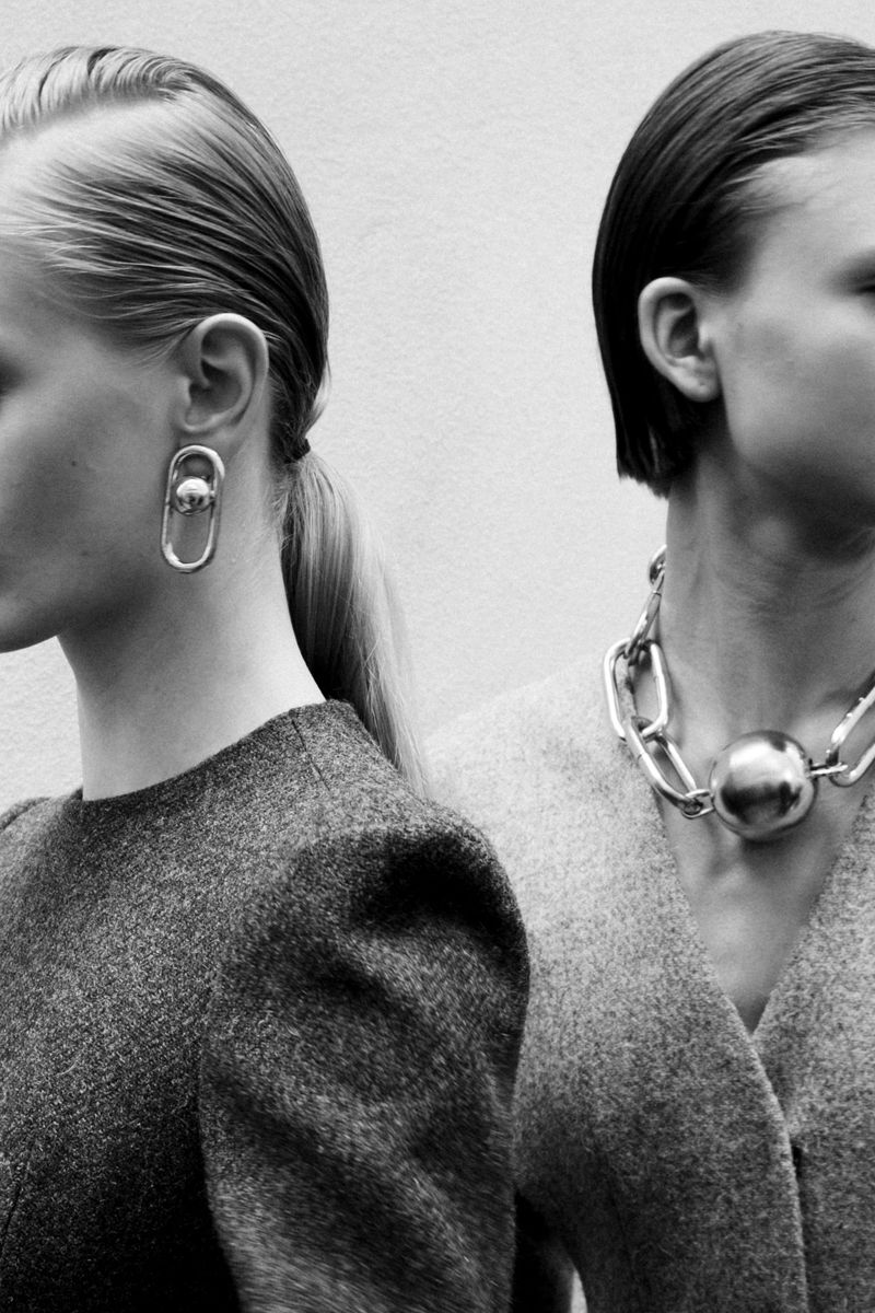 Two women pose with sleek hair, minimal makeup, and grey wool tops, showcasing silver chain necklaces and earrings against a neutral wall.