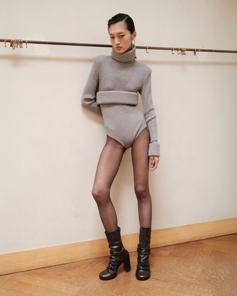 Model wearing a grey turtleneck knit bodysuit and dark boots posing against a plain wall backstage