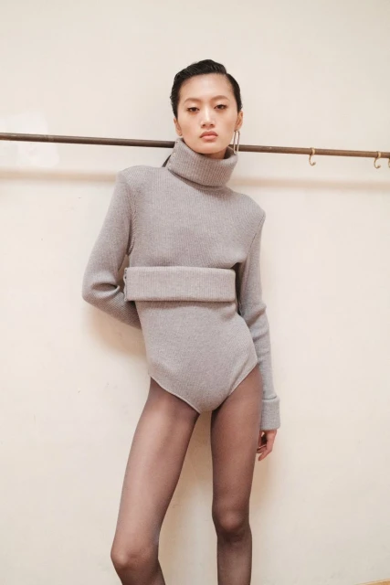 Asian fashion model poses in a light grey knit bodysuit with a high turtleneck, black tights, and large hoop earrings in a studio.