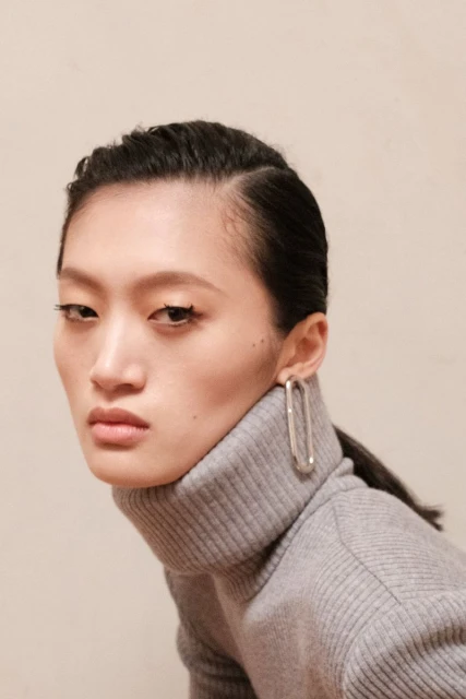 Close-up studio portrait of a pensive adult woman with dark hair, wearing a grey turtleneck and large, silver, oval-shaped earrings.