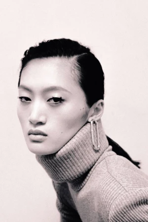 Close-up portrait of a model with dark hair wearing a ribbed turtleneck sweater and large metallic hoop earrings against a neutral studio backdrop