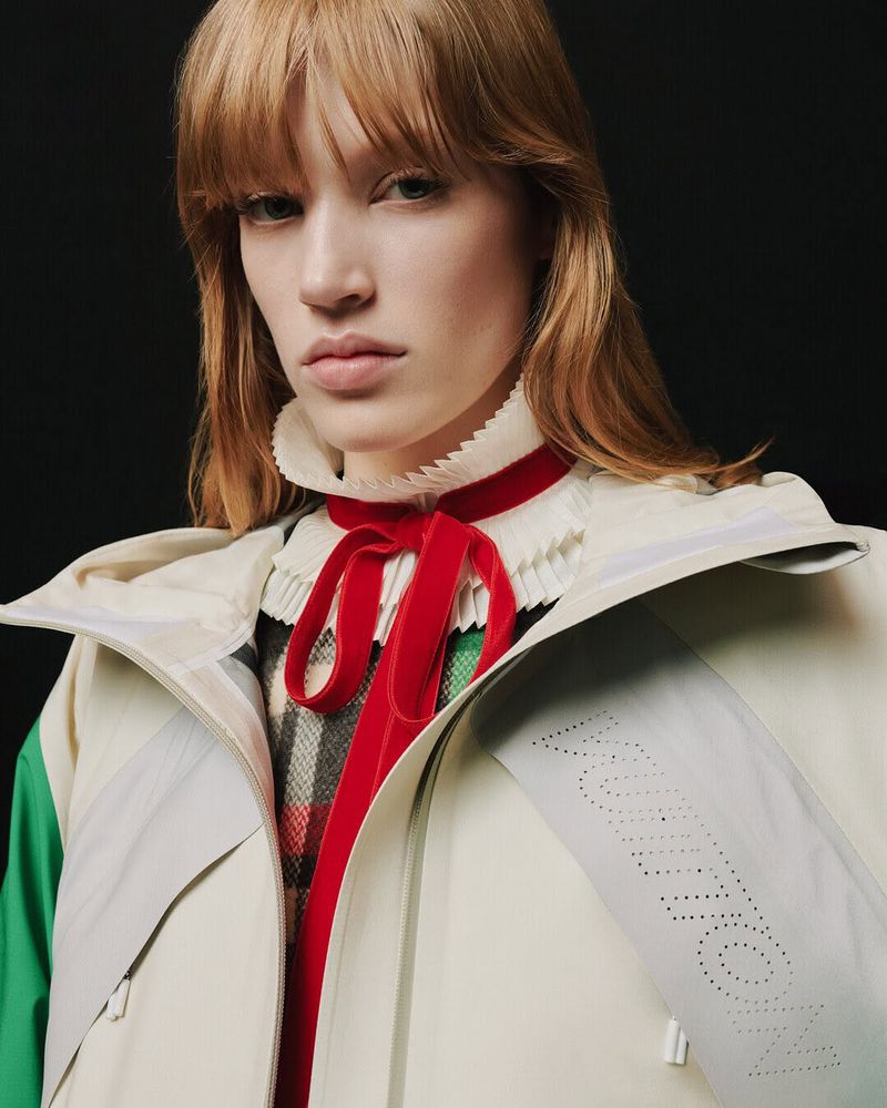 Close-up of a fair-skinned model with bangs, wearing a ruffled collar, red velvet ribbon, and a jacket with the word 'Vuitton' on the sleeve.