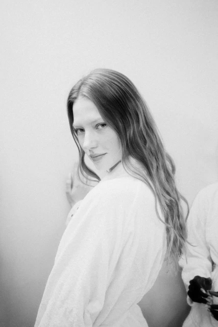 Monochrome portrait of a woman with long wavy hair, wearing a white shirt, looking over her shoulder indoors, against a plain background.
