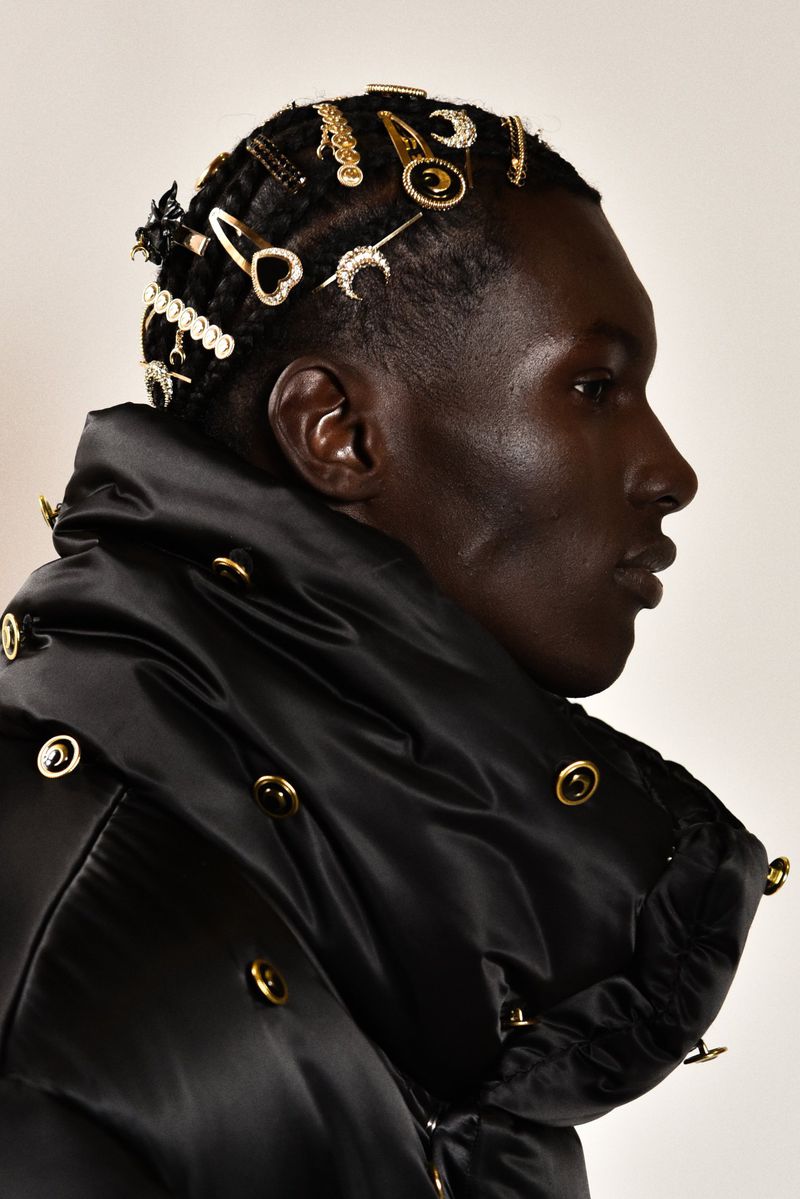 Close-up of a Black male model in a black puffer jacket decorated with gold accents, wearing hair clips, shot in a photo studio.