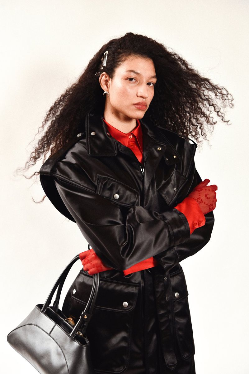 Woman with long curly hair wearing a black jacket and red top, holding a black purse, stands in a studio setting against a white background.