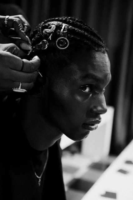 Close-up in black and white of a young adult man with braided hair being styled, hair clips are in his hair, rings on stylist's hand.