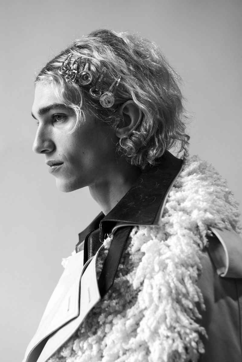 Black and white portrait of a young adult male in a fashionable coat, accented with hair clips and jewelry, looking off to the side.