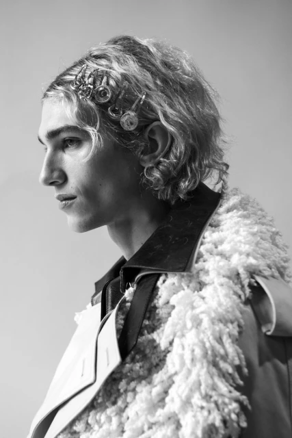 Black and white portrait of a young adult male in a fashionable coat, accented with hair clips and jewelry, looking off to the side.