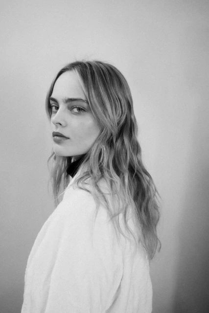 Monochrome portrait of a blonde woman with wavy hair looking over her shoulder wearing a white robe against a neutral backdrop.