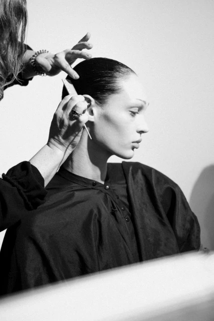 A fair-skinned person with dark hair is getting their hair styled by a person with bracelets in a monochrome, backlit indoor setting.